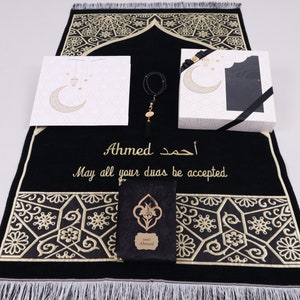 May include: A black prayer rug with gold embroidery and a personalized inscription that reads "Ahmed. May all your duas be accepted." The rug is accompanied by a white gift bag with a crescent moon and stars design, a black gift box with a black ribbon, and a prayer bead bracelet.