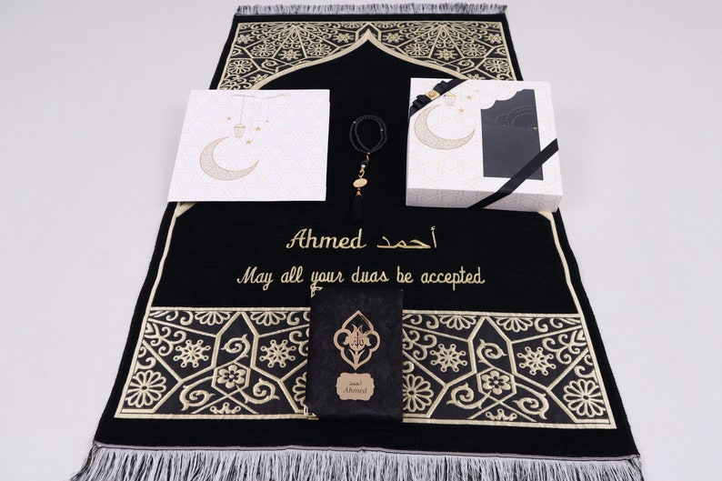 May include: A black prayer rug with gold embroidery and a personalized inscription that reads "Ahmed. May all your duas be accepted." The rug is accompanied by a white gift bag with a crescent moon and stars design, a black gift box with a black ribbon, and a prayer bead bracelet.