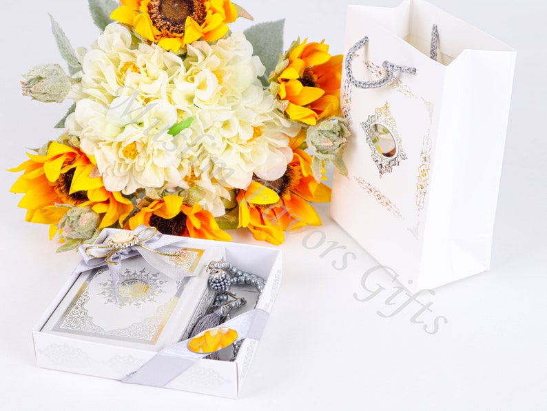 May include: A white gift box with a silver and gold design, containing a small book and a silver prayer beads. The box is surrounded by a bouquet of sunflowers and white hydrangeas.