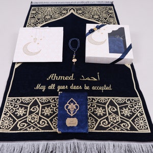Puede incluir: Una alfombra de oración azul con bordados dorados y una inscripción personalizada que dice "Ahmed. Que todas tus du'as sean aceptadas." La alfombra viene acompañada de un set de regalo que incluye un libro de oraciones azul, una bolsa de regalo blanca, un collar de cuentas de oración azul y una caja de regalo blanca con una bolsa de terciopelo azul.