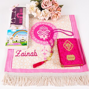 Peut inclure: Un tapis de pri&egrave;re rose avec un motif d&eacute;coratif, un Coran assorti et un chapelet de perles. Une bo&icirc;te cadeau avec une illustration d'arc-en-ciel et le nom "Zainab" est &eacute;galement incluse. Les articles sont dispos&eacute;s sur une surface blanche.