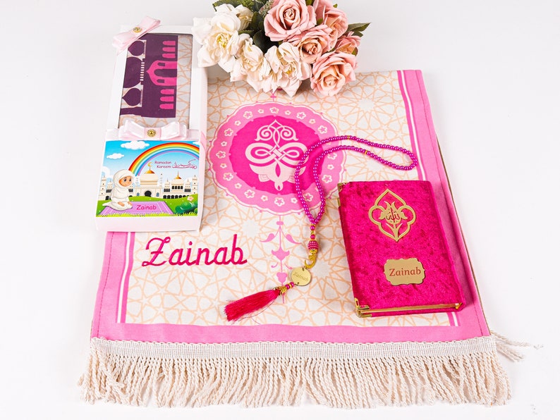 Peut inclure: Un tapis de pri&egrave;re rose avec un motif d&eacute;coratif, un Coran assorti et un chapelet de perles. Une bo&icirc;te cadeau avec une illustration d'arc-en-ciel et le nom "Zainab" est &eacute;galement incluse. Les articles sont dispos&eacute;s sur une surface blanche.