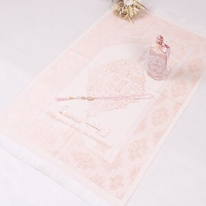 May include: A pink prayer rug with a white border and fringe. The rug features a floral design and Arabic calligraphy. A pink prayer bead set and a small pink gift box are on the rug.