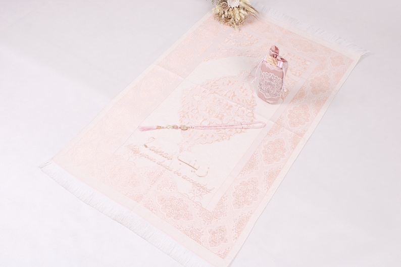 May include: A pink prayer rug with a white border and fringe. The rug features a floral design and Arabic calligraphy. A pink prayer bead set and a small pink gift box are on the rug.
