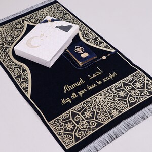 May include: A blue prayer rug with gold embroidery and a white box with a crescent moon design. The rug has the name "Ahmed" and the text "May all your duas be accepted" embroidered on it.