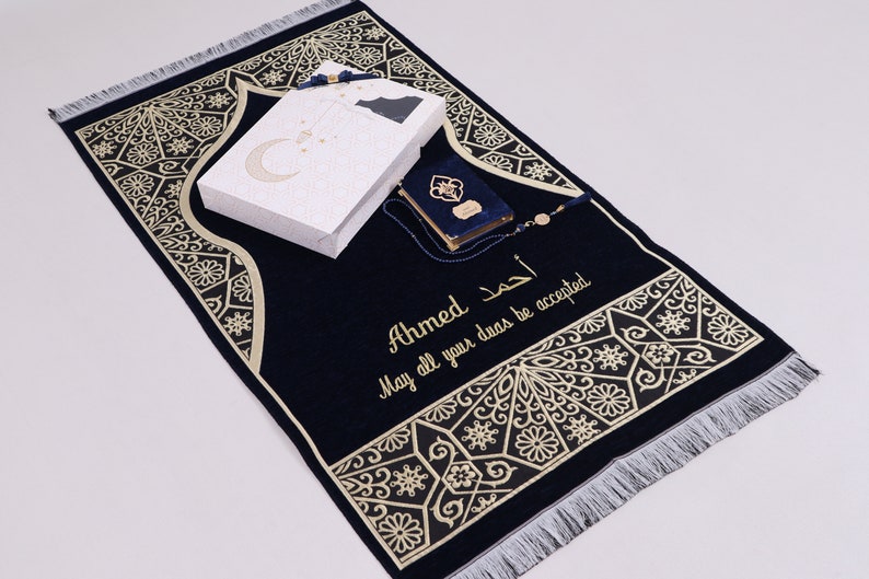 May include: A blue prayer rug with gold embroidery and a white box with a crescent moon design. The rug has the name "Ahmed" and the text "May all your duas be accepted" embroidered on it.
