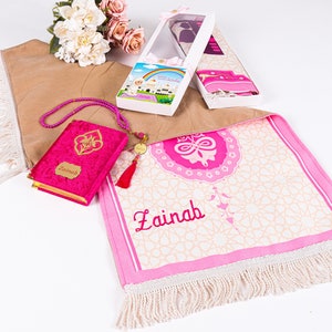 Peut inclure: Un tapis de pri&egrave;re rose avec le nom "Zainab" brod&eacute;, un Coran en velours rose et des accessoires de pri&egrave;re. Le tapis pr&eacute;sente un motif g&eacute;om&eacute;trique et une bordure &agrave; franges. Un coffret avec un n&oelig;ud est &eacute;galement visible.