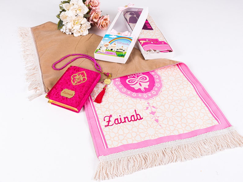 Peut inclure: Un tapis de pri&egrave;re rose avec le nom "Zainab" brod&eacute;, un Coran en velours rose et des accessoires de pri&egrave;re. Le tapis pr&eacute;sente un motif g&eacute;om&eacute;trique et une bordure &agrave; franges. Un coffret avec un n&oelig;ud est &eacute;galement visible.