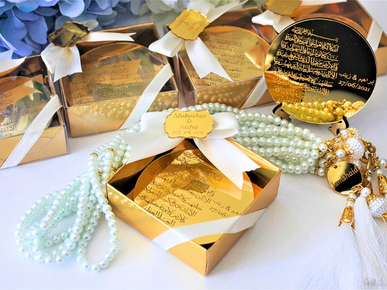 May include: Gold gift boxes with white ribbon and a small gold mirror with Arabic calligraphy. The boxes contain a white pearl prayer beads with a gold tassel.