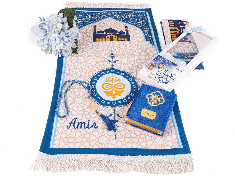 May include: An Islamic prayer mat with a mosque design, a blue beaded tasbih, a blue velvet Quran, and a gift box with the text "Eid Mubarak". The mat is beige with a blue border and fringe.