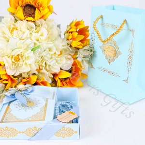 K&ouml;nnte beinhalten: Eine hellblaue Geschenkbox mit goldenen Verzierungen und einem goldenen Band. Die Box enth&auml;lt ein hellblaues Buch mit goldenen Lettern und hellblaue Gebetsperlen. Die Box steht auf einer wei&szlig;en Oberfl&auml;che neben einer hellblauen Geschenkt&uuml;te mit einem goldenen Henkel und goldenen Verzierungen.