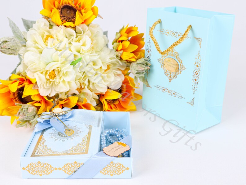 K&ouml;nnte beinhalten: Eine hellblaue Geschenkbox mit goldenen Verzierungen und einem goldenen Band. Die Box enth&auml;lt ein hellblaues Buch mit goldenen Lettern und hellblaue Gebetsperlen. Die Box steht auf einer wei&szlig;en Oberfl&auml;che neben einer hellblauen Geschenkt&uuml;te mit einem goldenen Henkel und goldenen Verzierungen.