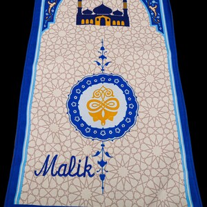 May include: A prayer rug with a beige geometric pattern and a blue border. The rug features a mosque illustration and the name "Malik" in script. The design includes a central medallion and decorative floral elements.