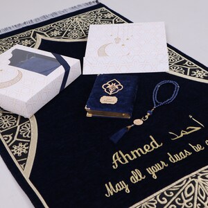 May include: A navy blue prayer rug with gold embroidery, a matching prayer book with a blue velvet cover, a set of blue prayer beads, and a white gift box with gold accents. The rug has the text "Ahmed May all your duas be accepted" embroidered on it.