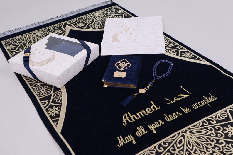 May include: A navy blue prayer rug with gold embroidery, a matching prayer book with a blue velvet cover, a set of blue prayer beads, and a white gift box with gold accents. The rug has the text "Ahmed May all your duas be accepted" embroidered on it.