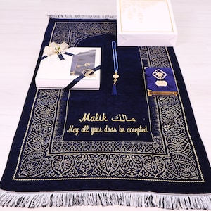 May include: A blue prayer rug with gold embroidery and a personalized inscription that reads "Malik  مالك May all your duas be accepted". The rug is part of a gift set that includes a white gift box, a blue prayer beads, and a small blue book.