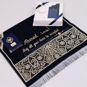 May include: A navy blue prayer rug with gold embroidery and a white fringe. The rug has a personalized inscription that reads "Ahmed May all your duas be accepted". The set includes a blue prayer book, a set of blue prayer beads, and a decorative box.