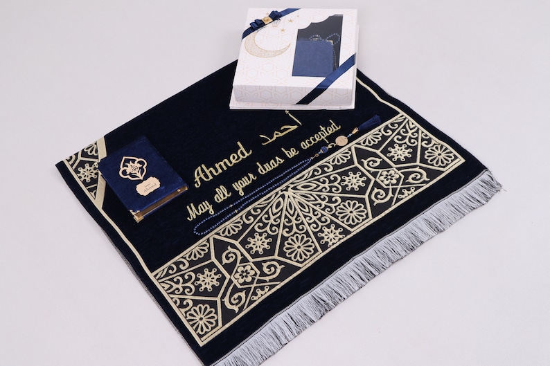 May include: A navy blue prayer rug with gold embroidery and a white fringe. The rug has a personalized inscription that reads "Ahmed May all your duas be accepted". The set includes a blue prayer book, a set of blue prayer beads, and a decorative box.