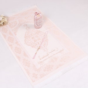 May include: A pink prayer rug with a white fringe and a floral design. The rug has a pink prayer bead necklace and a small pink box with a floral design on it. The text on the rug reads "Zikrullah - Keep your heart in remembrance."