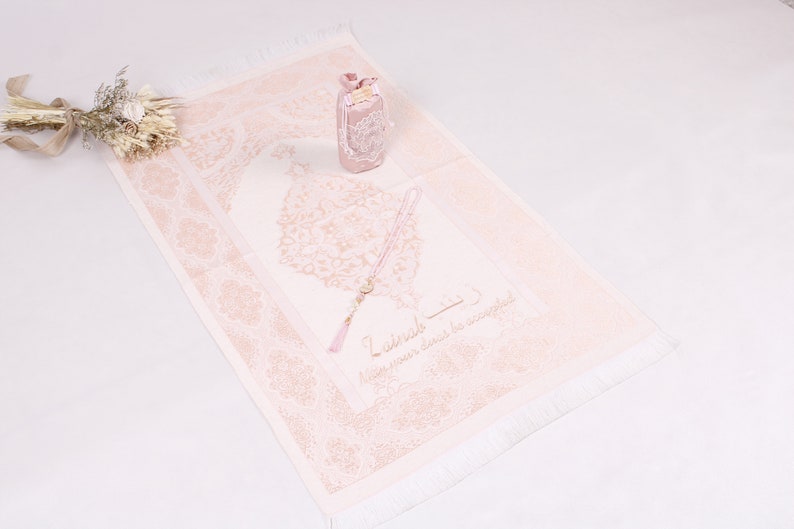 May include: A pink prayer rug with a white fringe and a floral design. The rug has a pink prayer bead necklace and a small pink box with a floral design on it. The text on the rug reads "Zikrullah - Keep your heart in remembrance."
