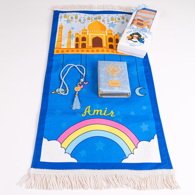 May include: A blue prayer rug with a rainbow and a mosque design, a rosary, and a small book. A box with the text "Eid Mubarak" is on the top right. The name "Amir" is written on the rug and the book.