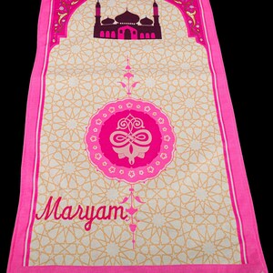 May include: A pink prayer rug with a mosque design at the top and the name "Maryam" in cursive. The rug has a beige background with a repeating star pattern and a pink border with fringe.