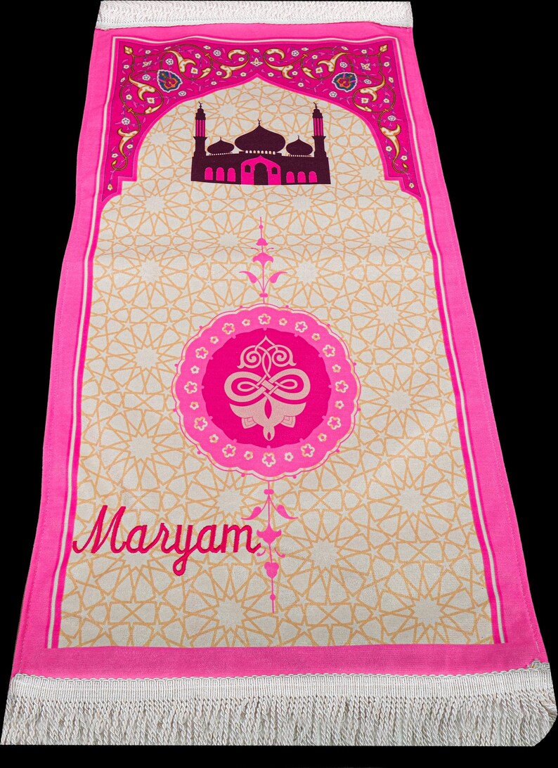 May include: A pink prayer rug with a mosque design at the top and the name "Maryam" in cursive. The rug has a beige background with a repeating star pattern and a pink border with fringe.