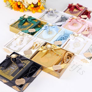 K&ouml;nnte beinhalten: Sortiment von Geschenkboxen mit Koranb&uuml;chern und Gebetsperlen. Die Boxen sind mit B&auml;ndern, Schleifen und goldenen Akzenten verziert. Die B&uuml;cher sind in verschiedenen Farben, darunter Gr&uuml;n, Blau, Wei&szlig; und Gold. Die Gebetsperlen sind wei&szlig;, schwarz und braun.