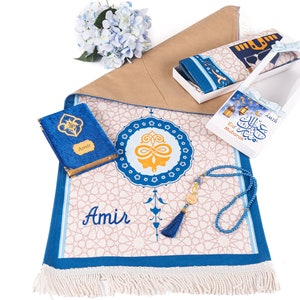 May include: A prayer mat with a blue and white geometric design, a blue velvet book, a box with a prayer rug, and a blue beaded tasbih. The mat has the name "Amir" on it. A gift set for religious occasions.
