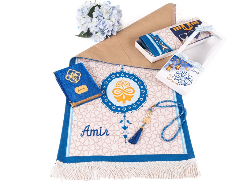 May include: A prayer mat with a blue and white geometric design, a blue velvet book, a box with a prayer rug, and a blue beaded tasbih. The mat has the name "Amir" on it. A gift set for religious occasions.