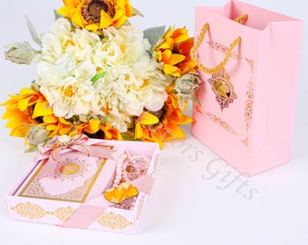 May include: A pink gift box with a gold trim and a pink ribbon. The box contains a pink Quran book, a set of prayer beads, and a small yellow flower. The box is sitting on a white surface next to a pink gift bag with a gold handle.