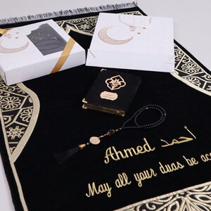 May include: A black prayer rug with gold embroidery and a decorative border. The rug is accompanied by a black prayer book, a set of prayer beads, and a gift box with a crescent moon design.