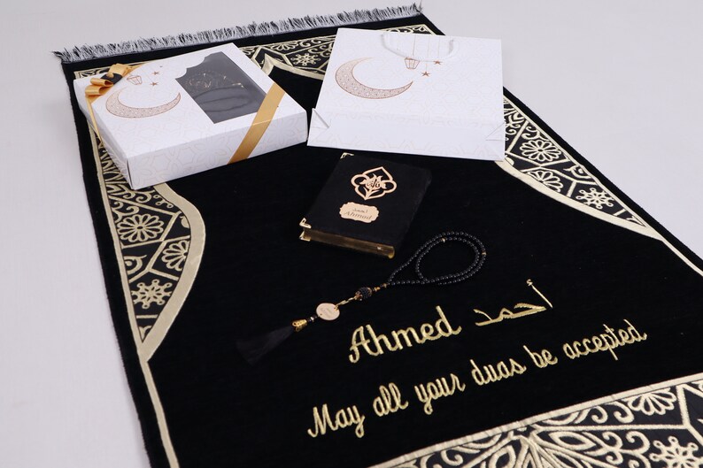 May include: A black prayer rug with gold embroidery and a decorative border. The rug is accompanied by a black prayer book, a set of prayer beads, and a gift box with a crescent moon design.