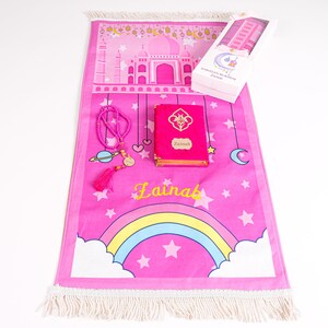 Peut inclure: Tapis de pri&egrave;re rose avec un arc-en-ciel, des &eacute;toiles et un motif de mosqu&eacute;e. Comprend un Coran rose, un chapelet et une bo&icirc;te cadeau. Le Coran est &eacute;tiquet&eacute; "Zainab" en lettres dor&eacute;es. Le tapis a une frange blanche.