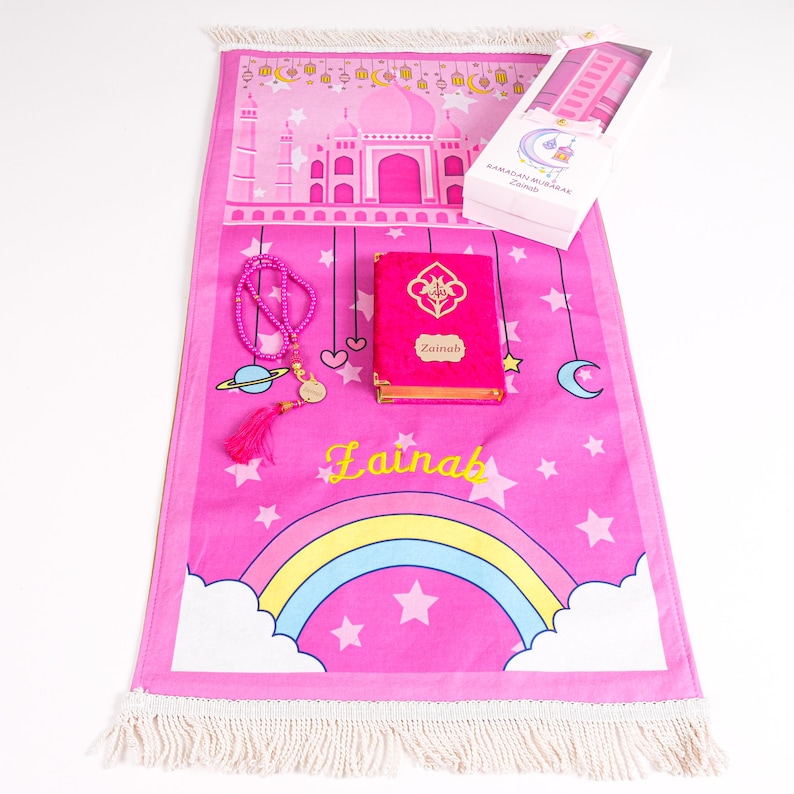 Peut inclure: Tapis de pri&egrave;re rose avec un arc-en-ciel, des &eacute;toiles et un motif de mosqu&eacute;e. Comprend un Coran rose, un chapelet et une bo&icirc;te cadeau. Le Coran est &eacute;tiquet&eacute; "Zainab" en lettres dor&eacute;es. Le tapis a une frange blanche.