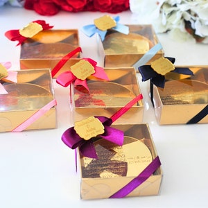 May include: Gold favor boxes with clear lids and personalized labels. Each box has a different colored ribbon and a gold-colored round disc inside. The labels have the names "Subhanahar & Aisha" and the date "July 27, 2023".