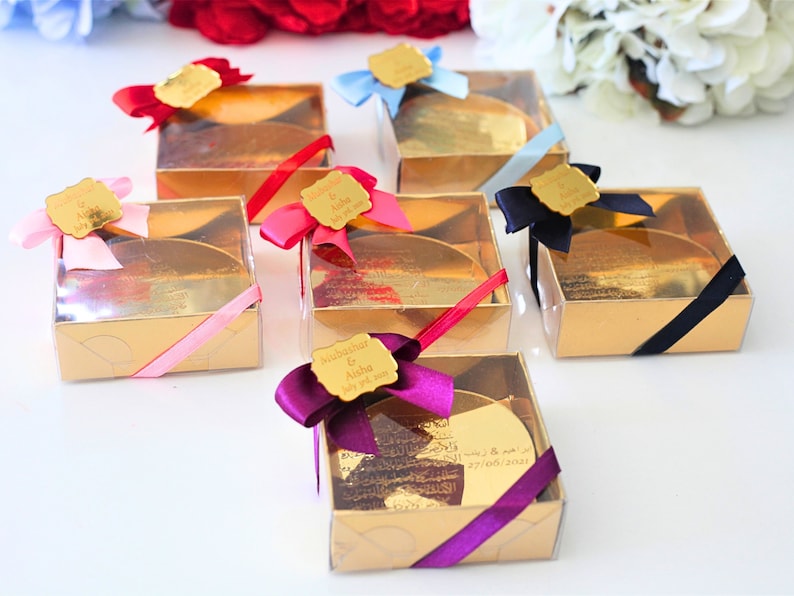 May include: Gold favor boxes with clear lids and personalized labels. Each box has a different colored ribbon and a gold-colored round disc inside. The labels have the names "Subhanahar & Aisha" and the date "July 27, 2023".