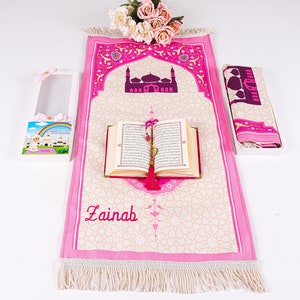 Peut inclure: Un tapis de pri&egrave;re rose avec un motif de mosqu&eacute;e, un Coran et une &eacute;charpe assortie. Le tapis est personnalis&eacute; avec le nom "Zainab". Une bo&icirc;te cadeau avec une illustration d'arc-en-ciel et de mosqu&eacute;e est &eacute;galement incluse. L'ensemble est con&ccedil;u pour la pri&egrave;re islamique.
