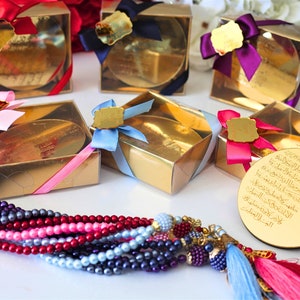 May include: Gold boxes with clear lids and colorful ribbons, containing round gold-colored plaques with Arabic calligraphy. The boxes are arranged on a white surface. A multi-colored beaded necklace with a tassel hangs in the foreground.
