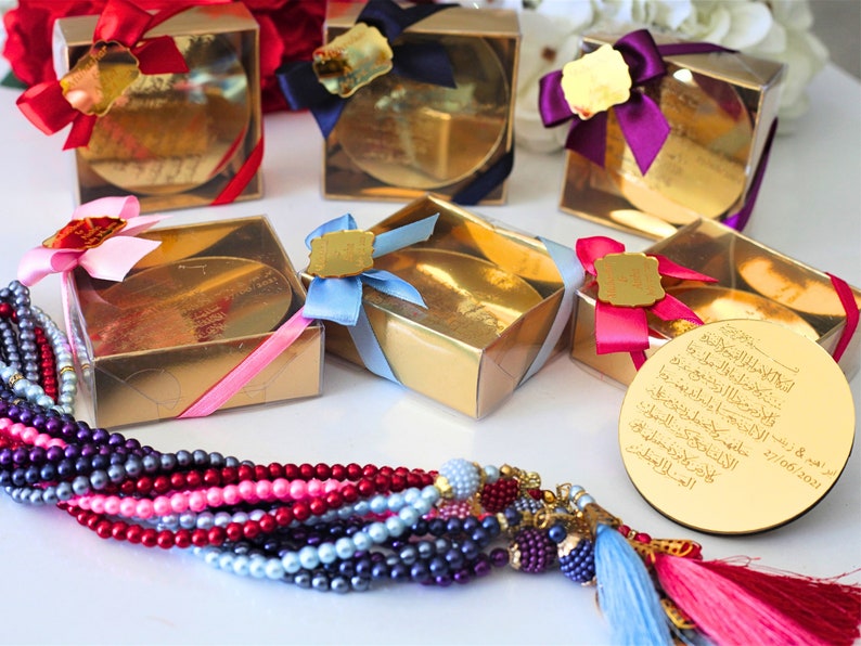 May include: Gold boxes with clear lids and colorful ribbons, containing round gold-colored plaques with Arabic calligraphy. The boxes are arranged on a white surface. A multi-colored beaded necklace with a tassel hangs in the foreground.