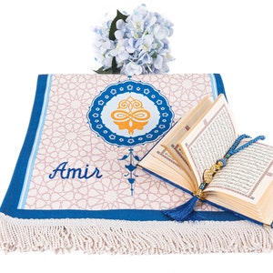 May include: A prayer rug with a geometric pattern and the name "Amir" in script. An open book with gold-edged pages and a blue tassel is next to the rug. A blue beaded rosary and a bouquet of blue flowers complete the scene.