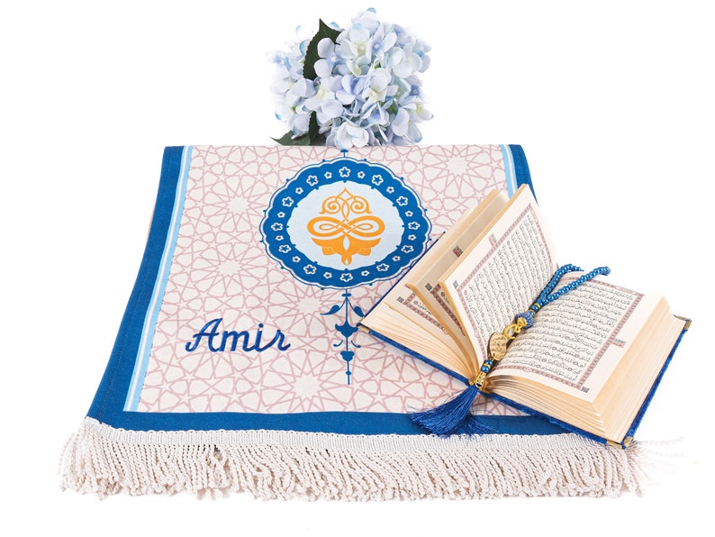 May include: A prayer rug with a geometric pattern and the name "Amir" in script. An open book with gold-edged pages and a blue tassel is next to the rug. A blue beaded rosary and a bouquet of blue flowers complete the scene.