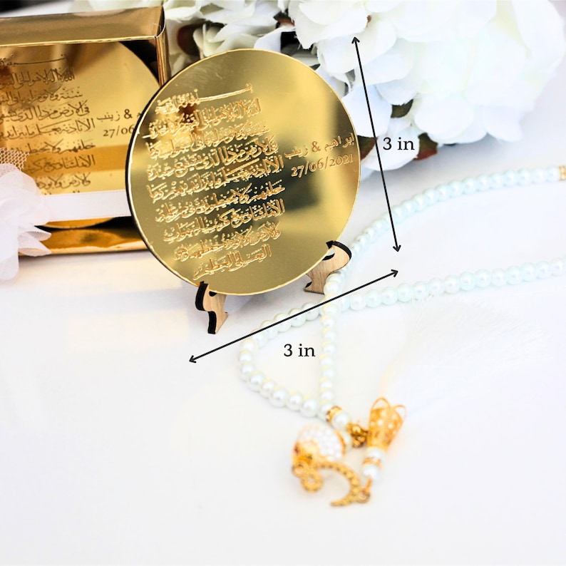 May include: A gold-colored round mirror with Arabic calligraphy and a date inscribed on it. The mirror is on a small wooden stand and measures 3 inches in diameter.