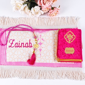 Peut inclure: Un tapis de pri&egrave;re rose, un Coran assorti et un chapelet de perles sont pr&eacute;sent&eacute;s. Le tapis et le Coran sont personnalis&eacute;s avec le nom "Zainab". Le Coran a des accents dor&eacute;s et un fermoir en m&eacute;tal dor&eacute;. Un pompon rose est suspendu au chapelet.
