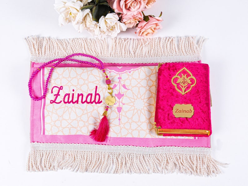 Peut inclure: Un tapis de pri&egrave;re rose, un Coran assorti et un chapelet de perles sont pr&eacute;sent&eacute;s. Le tapis et le Coran sont personnalis&eacute;s avec le nom "Zainab". Le Coran a des accents dor&eacute;s et un fermoir en m&eacute;tal dor&eacute;. Un pompon rose est suspendu au chapelet.