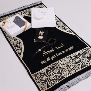 May include: A black prayer rug with gold embroidery and a personalized message that reads "Ahmed May all your duas be accepted". The rug is accompanied by a small black book, a white gift bag with a crescent moon design, and a set of prayer beads.