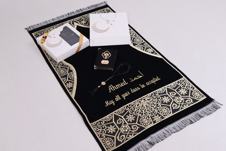 May include: A black prayer rug with gold embroidery and a personalized message that reads "Ahmed May all your duas be accepted". The rug is accompanied by a small black book, a white gift bag with a crescent moon design, and a set of prayer beads.