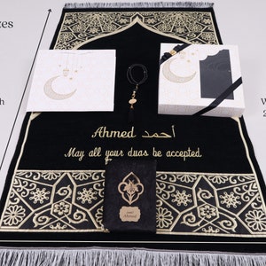 May include: A black prayer rug with gold embroidery and a personalized inscription that reads "Ahmed أحمد May all your duas be accepted". The rug is 51 inches long and 27 inches wide. It comes with a matching prayer mat, a small gift box, and a prayer bead bracelet.