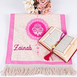 Peut inclure: Un tapis de pri&egrave;re rose avec un motif g&eacute;om&eacute;trique et le nom "Zainab" brod&eacute; en rouge. Un livre ouvert avec des pages dor&eacute;es et un chapelet de perles roses reposent sur le tapis. Un bouquet de fleurs roses et blanches est en arri&egrave;re-plan.