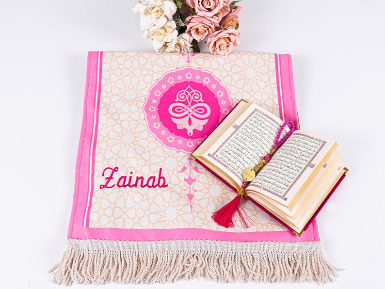 Peut inclure: Un tapis de pri&egrave;re rose avec un motif g&eacute;om&eacute;trique et le nom "Zainab" brod&eacute; en rouge. Un livre ouvert avec des pages dor&eacute;es et un chapelet de perles roses reposent sur le tapis. Un bouquet de fleurs roses et blanches est en arri&egrave;re-plan.
